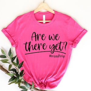 Op de afbeelding: Roze T-shirt met zwarte tekst die "Are we there yet? #roadtrip" zegt