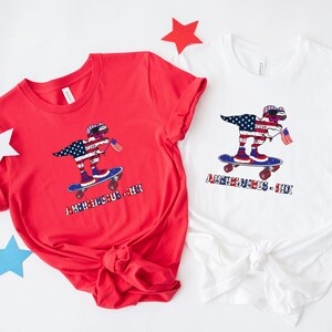 Puede incluir: Dos camisetas con un gráfico de un dinosaurio con gafas de sol y un pañuelo rojo, blanco y azul, montando un monopatín. El dinosaurio sostiene una pequeña bandera estadounidense. El texto "Amerisaurus - Rex" está impreso debajo del dinosaurio. Una camiseta es roja y la otra es blanca.