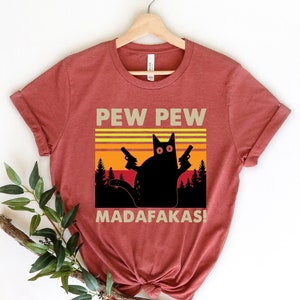 May include: Red t-shirt with a black cat holding two guns and a retro sunset graphic. The text "PEW PEW MADAFAKAS!" is printed on the shirt.