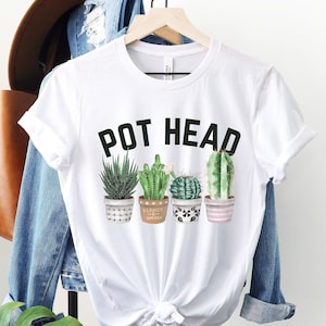 May include: White t-shirt with the text "POT HEAD" in black letters and a row of four potted cactus plants. The cactus plants are green and brown with white pots.