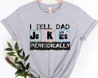 Puede incluir: Camiseta gris con el texto "I tell dad jokes periodically" y un gráfico de los elementos químicos para "Jo", "K" y "Es" con sus números atómicos.
