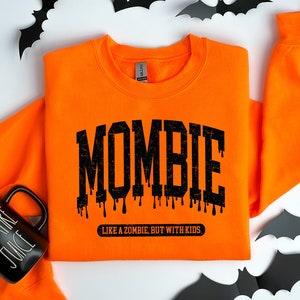 May include: Orange sweatshirt with black text that reads "Mombie: Like a zombie, but with kids". The sweatshirt is laid out on a white surface with black bats scattered around it. A black mug with white text that reads "Vampire Juice" is also in the image.