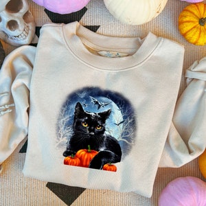 May include: A beige sweatshirt with a black cat and pumpkins in front of a full moon. The cat has yellow eyes and is looking at the viewer. The moon is surrounded by bats.