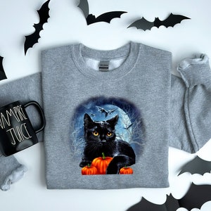 May include: A gray sweatshirt with a black cat sitting on a pile of orange pumpkins in front of a full moon and bats. The cat has yellow eyes and is looking at the viewer.