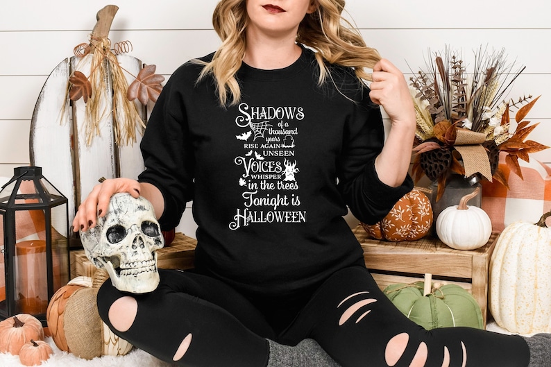 K&ouml;nnte beinhalten: Ein schwarzes Sweatshirt mit einem wei&szlig;en Grafikdruck, der "Shadows of a thousand years rise again unseen voices whisper in the trees tonight is Halloween" lautet.