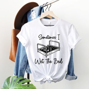 K&ouml;nnte beinhalten: Wei&szlig;es T-Shirt mit einer schwarzen Grafik eines Bettes, in dem Pflanzen wachsen. Der Text "Sometimes I Wet the Bed" ist unter der Grafik gedruckt.