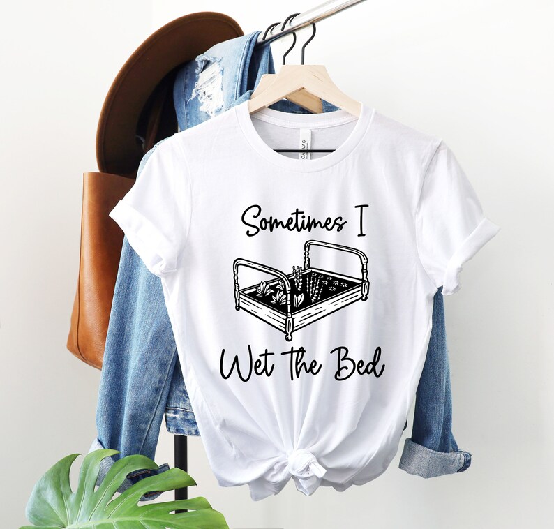 K&ouml;nnte beinhalten: Wei&szlig;es T-Shirt mit einer schwarzen Grafik eines Bettes, in dem Pflanzen wachsen. Der Text "Sometimes I Wet the Bed" ist unter der Grafik gedruckt.