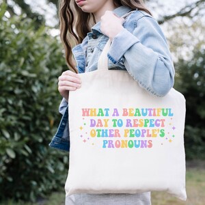 May include: A white tote bag with the text "What a beautiful day to respect other people's pronouns" in rainbow colors. The text is in a retro font style.