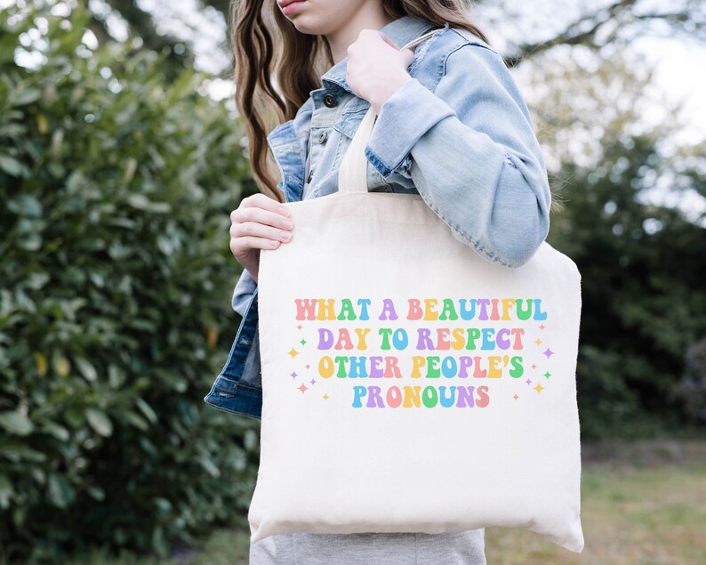 May include: A white tote bag with the text "What a beautiful day to respect other people's pronouns" in rainbow colors. The text is in a retro font style.