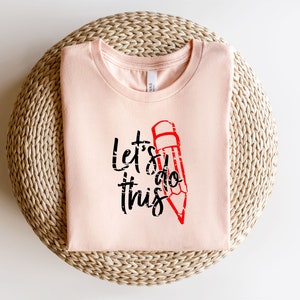 K&ouml;nnte beinhalten: Hellrosa T-Shirt mit dem Text "Let's do this" in Schwarz und einer roten Bleistiftgrafik. Das gefaltete Shirt liegt auf einer gewebten, kreisf&ouml;rmigen Oberfl&auml;che.