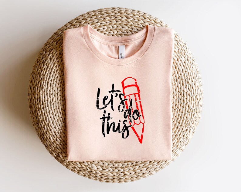 K&ouml;nnte beinhalten: Hellrosa T-Shirt mit dem Text "Let's do this" in Schwarz und einer roten Bleistiftgrafik. Das gefaltete Shirt liegt auf einer gewebten, kreisf&ouml;rmigen Oberfl&auml;che.