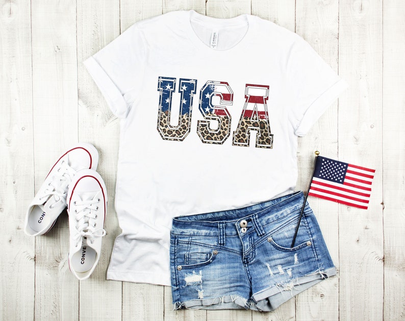 May include: White t-shirt with a graphic of the word "USA" in red, white, and blue with a leopard print background. The shirt is paired with denim shorts and white sneakers.