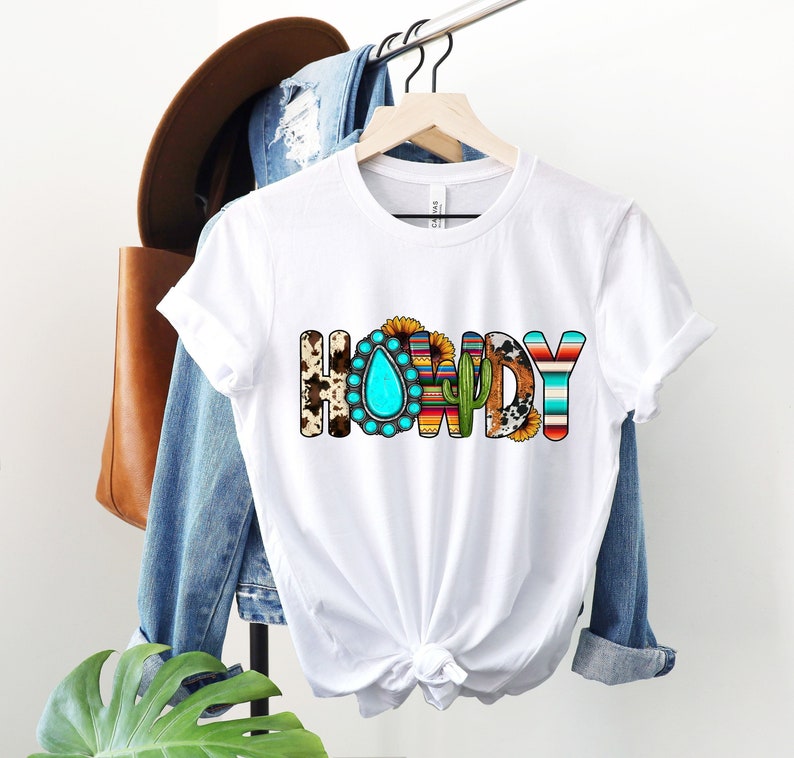 K&ouml;nnte beinhalten: Wei&szlig;es T-Shirt mit dem Wort "HOWDY" in bunten Buchstaben. Die Buchstaben sind mit s&uuml;dwestlichen Designs verziert, darunter T&uuml;rkis, Sonnenblumen, Kakteen und Kuhfell.