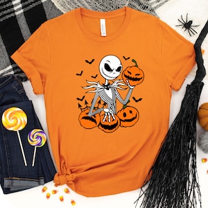 May include: Orange t-shirt with a black and white graphic of Jack Skellington from The Nightmare Before Christmas holding a pumpkin. The graphic also includes bats and pumpkins.