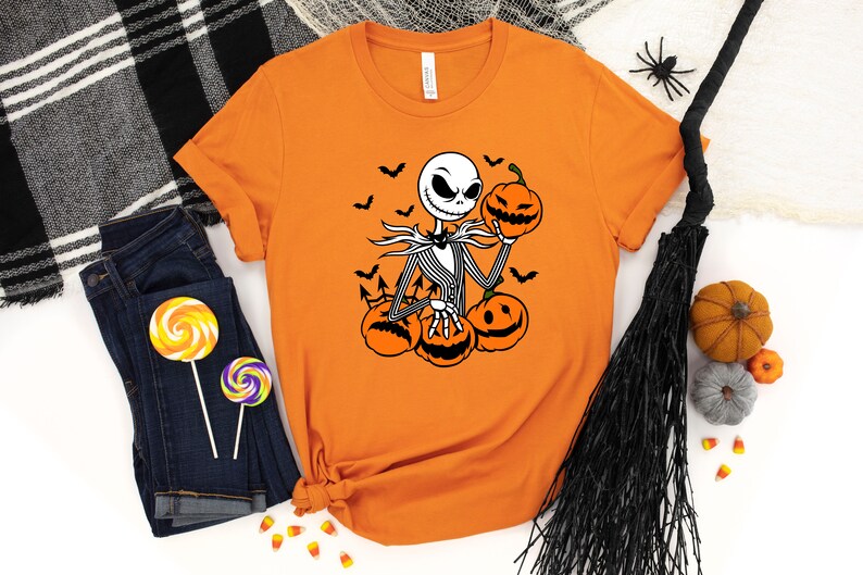 May include: Orange t-shirt with a black and white graphic of Jack Skellington from The Nightmare Before Christmas holding a pumpkin. The graphic also includes bats and pumpkins.