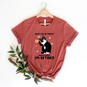 以下が含まれることがあります： コーヒーカップを持った白黒の猫が描かれた赤いTシャツ。Tシャツには「今日は何曜日？誰が気にする？私は引退した。」という文字が書かれています。