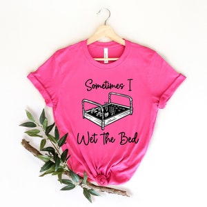 K&ouml;nnte beinhalten: Pinkes T-Shirt mit einer schwarzen Grafik eines Bettes mit Pflanzen, die darin wachsen, und dem Text "Sometimes I Wet the Bed".