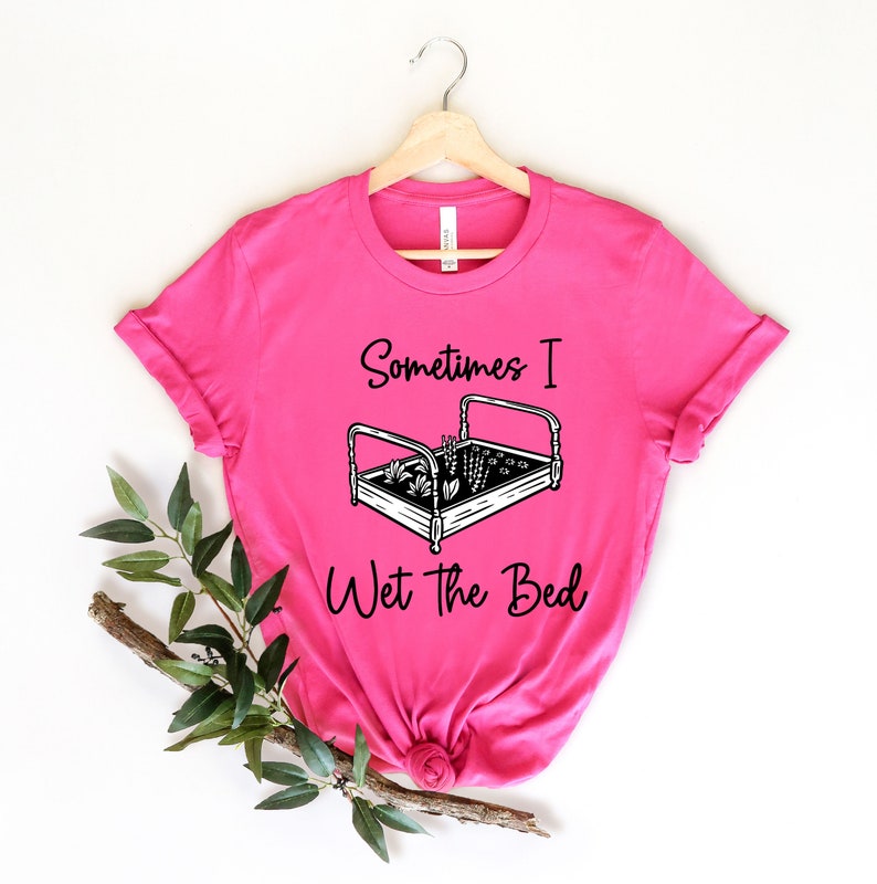 K&ouml;nnte beinhalten: Pinkes T-Shirt mit einer schwarzen Grafik eines Bettes mit Pflanzen, die darin wachsen, und dem Text "Sometimes I Wet the Bed".