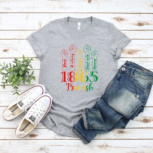 May include: Gray t-shirt with a graphic design of five raised fists with text that reads "Black History", "Stand with Slavery", and "1865 Free-ish".