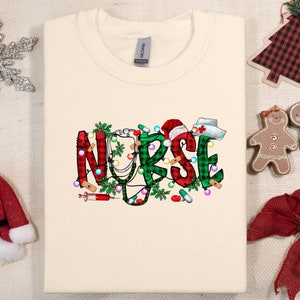 May include: A cream-colored t-shirt with a Christmas-themed design featuring the word "NURSE" in green, red, and white plaid with a stethoscope, candy canes, and Christmas lights.