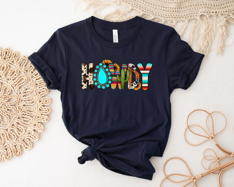 K&ouml;nnte beinhalten: Ein marineblaues T-Shirt mit dem Wort "Howdy" in bunten Buchstaben. Die Buchstaben sind mit s&uuml;dwestlich inspirierten Mustern verziert, darunter ein t&uuml;rkisfarbener Tropfen, ein Kaktus und eine Sonnenblume.