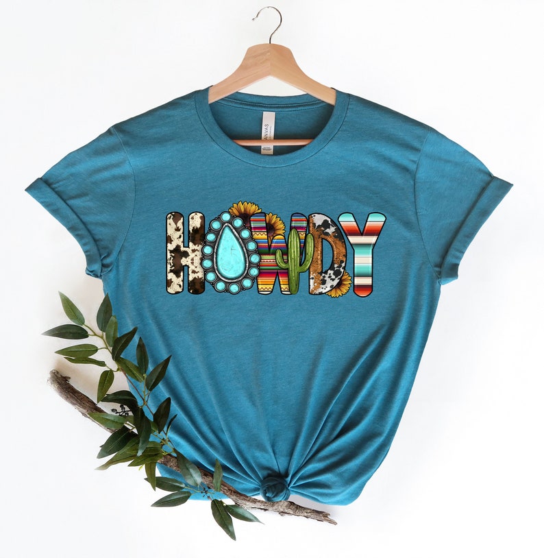 K&ouml;nnte beinhalten: Tealblaues T-Shirt mit dem Wort "HOWDY" in bunten Buchstaben. Die Buchstaben sind mit s&uuml;dwestlichen Mustern, T&uuml;rkis-Steinen, Kakteen und Sonnenblumen verziert.