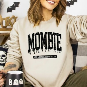 May include: A beige sweatshirt with a black graphic that says "Mombie" with dripping black paint and the text "Like a zombie, but with kids".