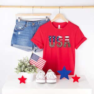 May include: Red t-shirt with the word "USA" printed on it in white letters with a red, white, and blue American flag and leopard print design. The shirt is on a hanger with a pair of blue denim shorts and a small American flag.