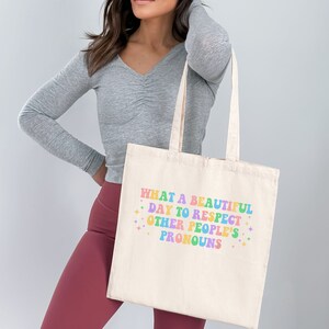 May include: A white canvas tote bag with the text "WHAT A BEAUTIFUL DAY TO RESPECT OTHER PEOPLE'S PRONOUNS" printed in rainbow colors.