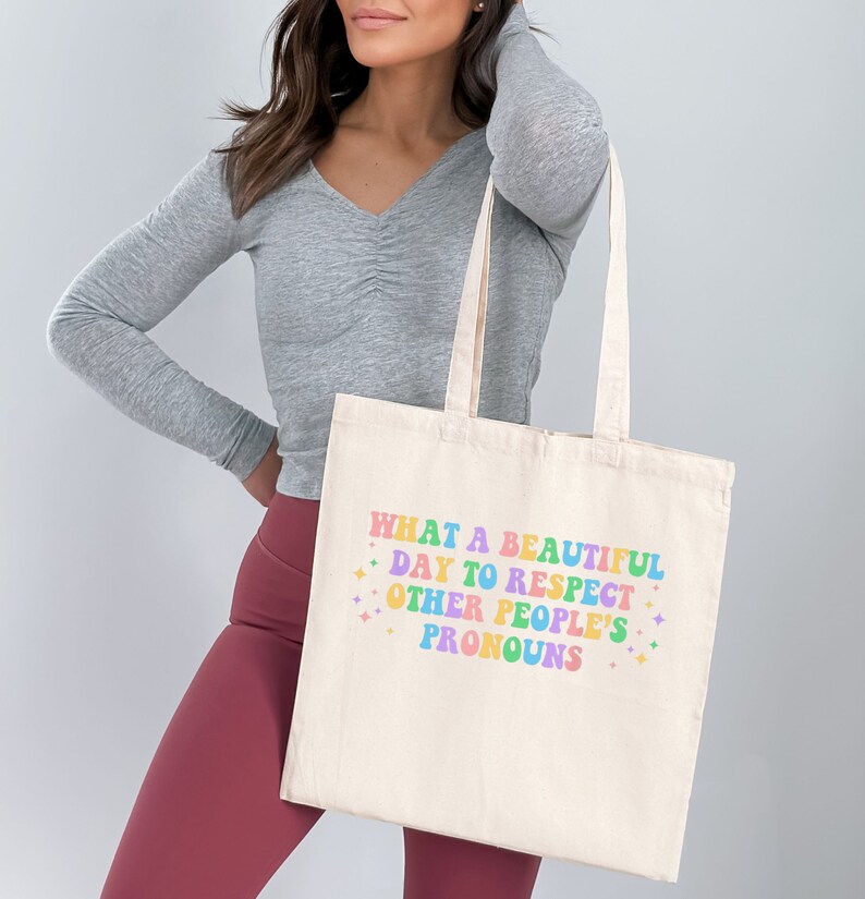 May include: A white canvas tote bag with the text "WHAT A BEAUTIFUL DAY TO RESPECT OTHER PEOPLE'S PRONOUNS" printed in rainbow colors.