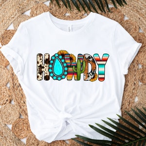 K&ouml;nnte beinhalten: Wei&szlig;es T-Shirt mit dem Wort "HOWDY" in bunten Buchstaben. Die Buchstaben sind mit s&uuml;dwestlichen Mustern verziert, darunter t&uuml;rkisfarbene Steine, Kakteen und Sonnenblumen.