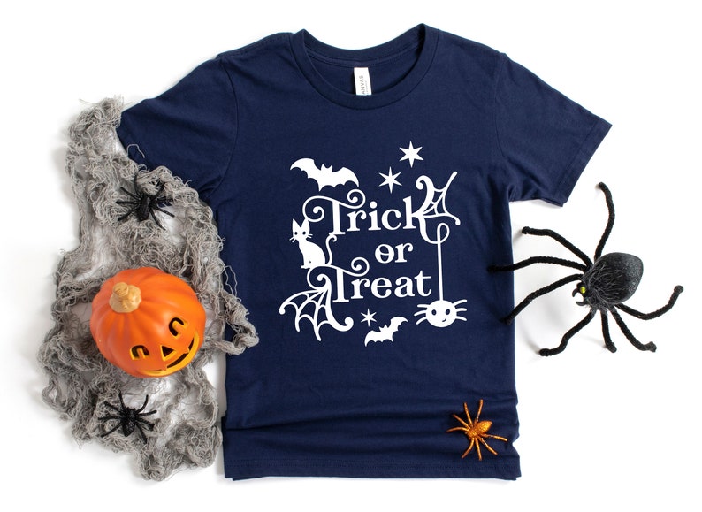 May include: A navy blue t-shirt with a white graphic design that says "Trick or Treat" with a spider, bat, and cat. The design is surrounded by stars and spiderwebs.