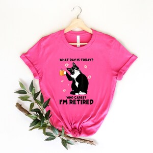 以下が含まれることがあります： ピンクのTシャツに、コーヒーカップを持った白黒の猫が描かれています。Tシャツには「今日は何曜日？誰が気にする？私は引退した。」という文字が書かれています。