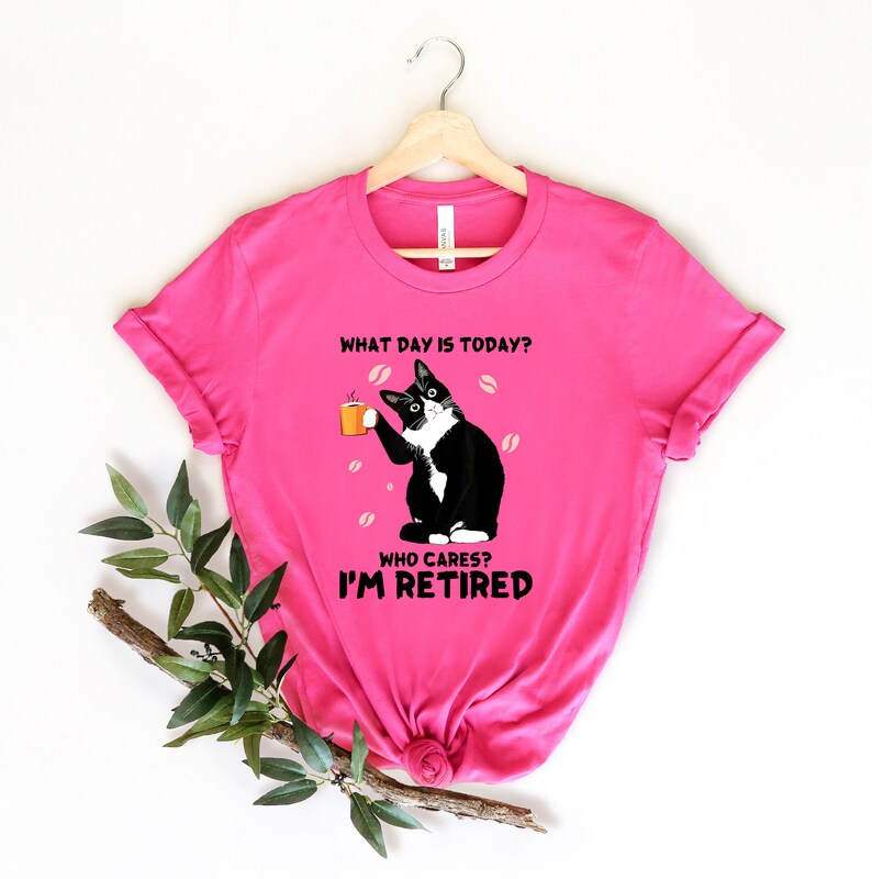 以下が含まれることがあります： ピンクのTシャツに、コーヒーカップを持った白黒の猫が描かれています。Tシャツには「今日は何曜日？誰が気にする？私は引退した。」という文字が書かれています。