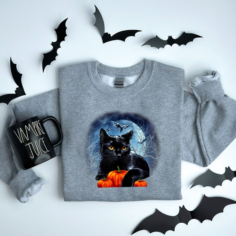 May include: A gray sweatshirt with a black cat sitting on a pile of orange pumpkins in front of a full moon and bats. The cat has yellow eyes and is looking at the viewer.
