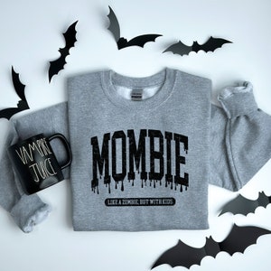 May include: A gray sweatshirt with the text "MOMBIE" in black dripping letters. The text "Like a zombie, but with kids" is printed below the word "MOMBIE".