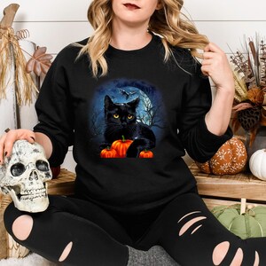 May include: Black cat with yellow eyes sitting on a pile of orange pumpkins in front of a full moon. The cat is surrounded by a dark forest with trees and a bat flying overhead. The image is on a black t-shirt.