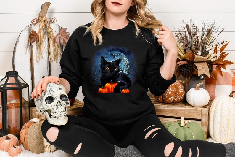 May include: Black cat with yellow eyes sitting on a pile of orange pumpkins in front of a full moon. The cat is surrounded by a dark forest with trees and a bat flying overhead. The image is on a black t-shirt.