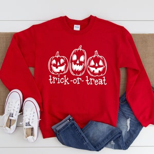 Może przedstawiać: Czerwona bluza z białym nadrukiem trzech dyni z wyciętymi twarzami i napisem "trick-or-treat". Obraz zawiera niebieskie dżinsy i białe trampki, idealne na Halloween.