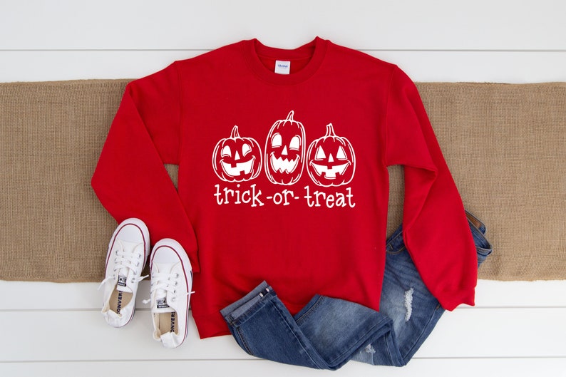 Może przedstawiać: Czerwona bluza z białym nadrukiem trzech dyni z wyciętymi twarzami i napisem "trick-or-treat". Obraz zawiera niebieskie dżinsy i białe trampki, idealne na Halloween.