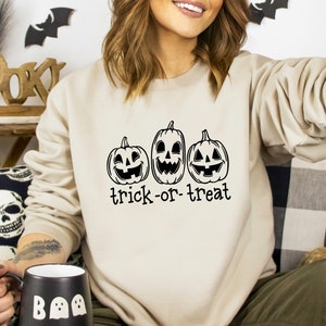Może przedstawiać: Beżowa bluza na Halloween z trzema uśmiechniętymi dyniami w czarnym tuszu. Pod dyniami nadrukowano napis "trick-or-treat". Bluza ma neutralny kolor, idealny na sezon jesienny.