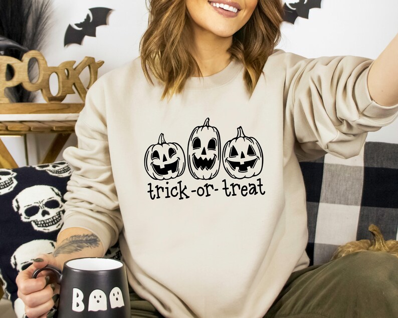 Może przedstawiać: Beżowa bluza na Halloween z trzema uśmiechniętymi dyniami w czarnym tuszu. Pod dyniami nadrukowano napis "trick-or-treat". Bluza ma neutralny kolor, idealny na sezon jesienny.