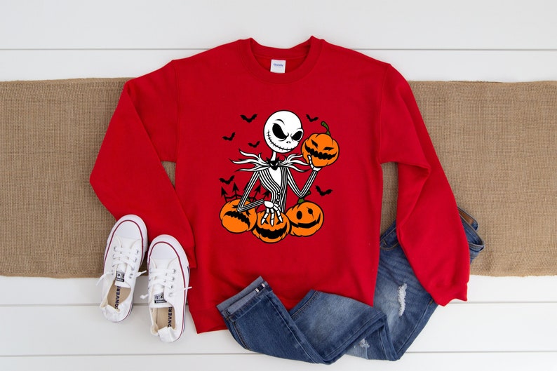 May include: Red sweatshirt with a black and white illustration of Jack Skellington from The Nightmare Before Christmas holding a pumpkin. The illustration also includes bats and pumpkins.
