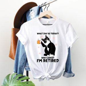 以下が含まれることがあります： コーヒーカップを持った白黒の猫が描かれた白いTシャツ。シャツのテキストは「今日は何曜日？誰が気にする？私は引退した。」