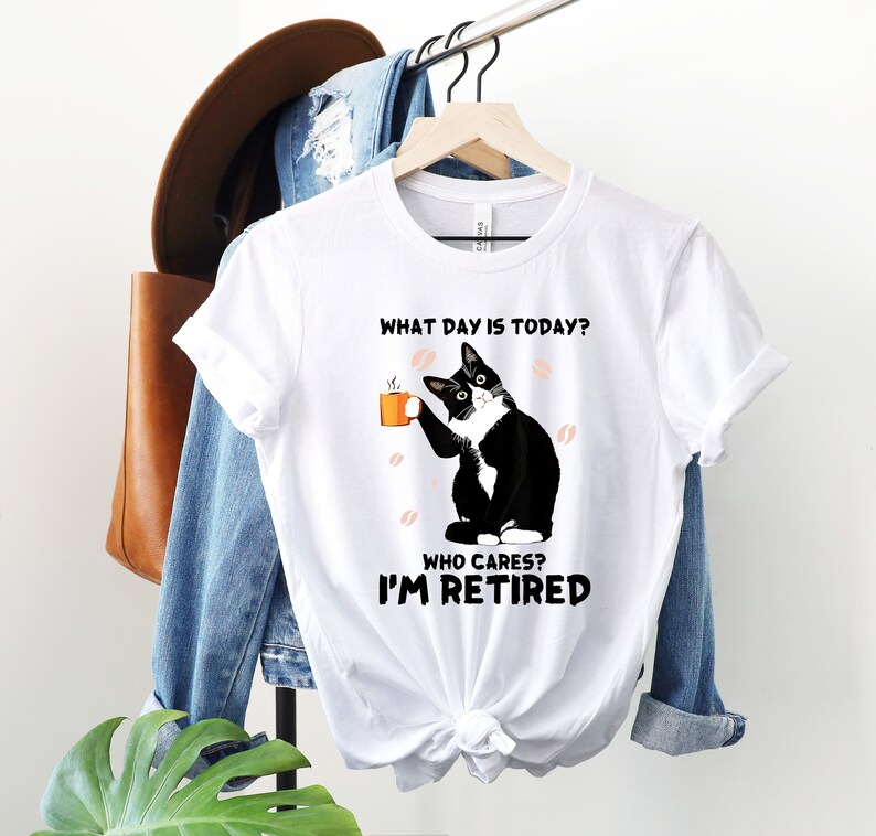 以下が含まれることがあります： コーヒーカップを持った白黒の猫が描かれた白いTシャツ。シャツのテキストは「今日は何曜日？誰が気にする？私は引退した。」