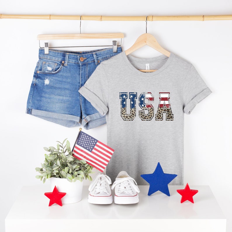 May include: A grey t-shirt with the word "USA" printed on it in large, bold letters. The letters are decorated with an American flag pattern and a leopard print. The shirt is hanging on a wooden hanger.