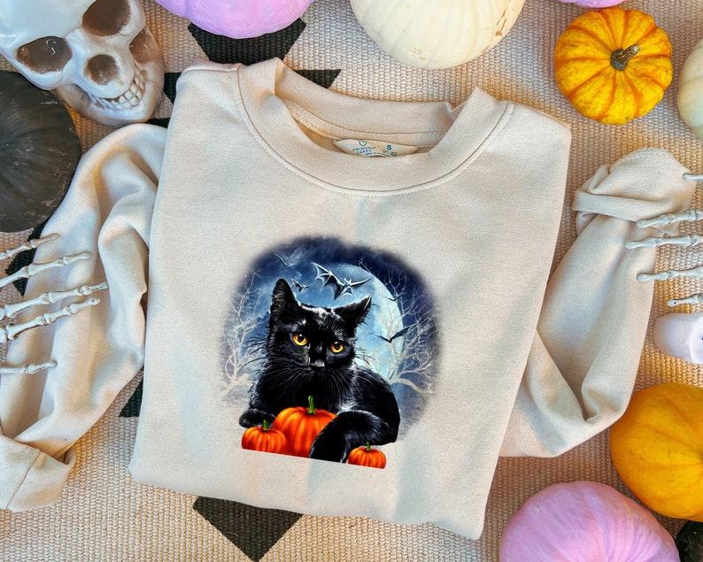 May include: A beige sweatshirt with a black cat and pumpkins in front of a full moon. The cat has yellow eyes and is looking at the viewer. The moon is surrounded by bats.