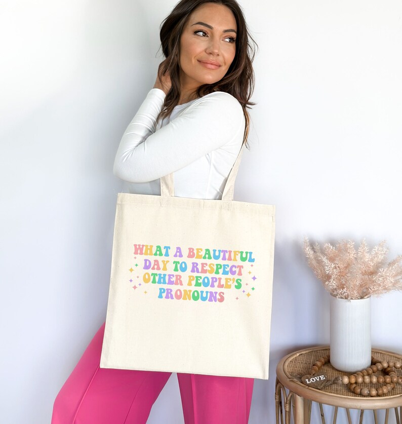 May include: A white tote bag with the text "WHAT A BEAUTIFUL DAY TO RESPECT OTHER PEOPLE'S PRONOUNS" in rainbow colors.