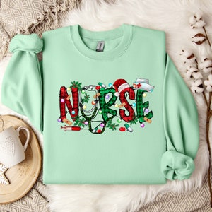 May include: A light green sweatshirt with a Christmas-themed design that says "NURSE" in red and green plaid with a stethoscope, a syringe, and other medical symbols.