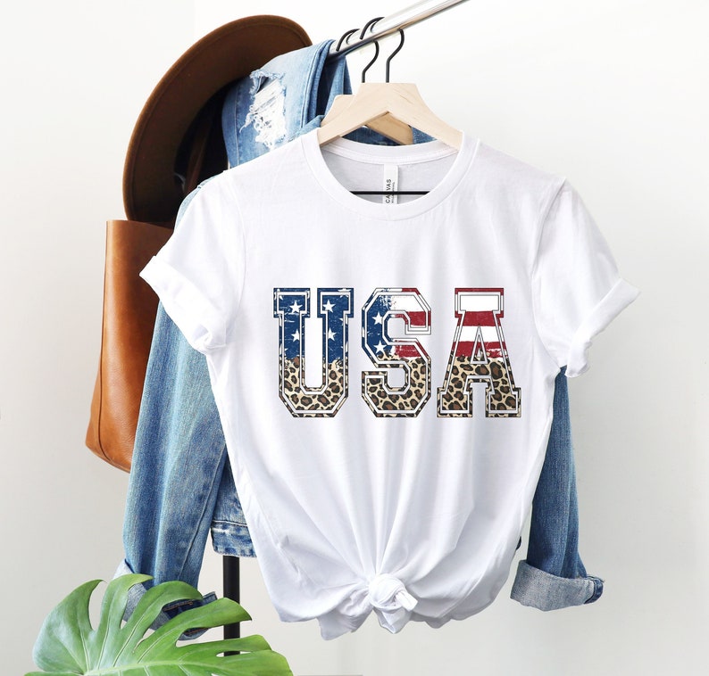 May include: White t-shirt with a distressed graphic of the word "USA" in bold letters. The letters are filled with a red, white, and blue American flag pattern and a leopard print.